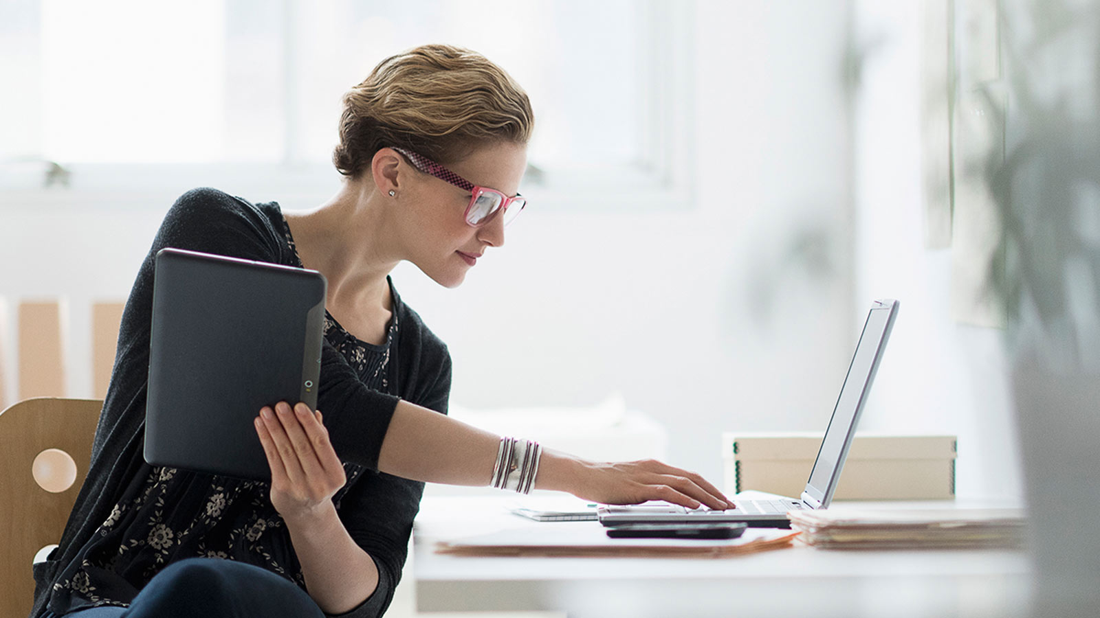 Woman seated at desk using laptop and holding a tablet with a smart phone nearby.