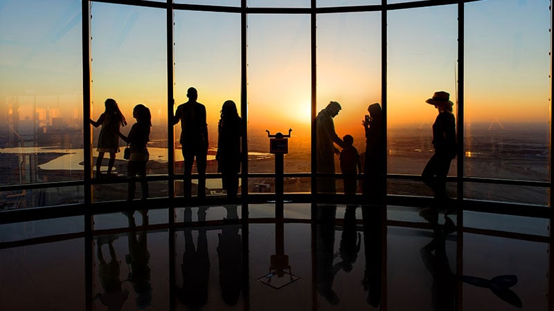 Silhouettes of people stand by large observation windows at sunset, overlooking a cityscape and water below.