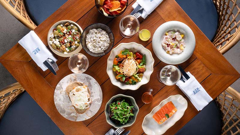 A wooden table set with various dishes including salads, bread, salmon, dip, vegetable bowls, drinks, and utensils, viewed from above.