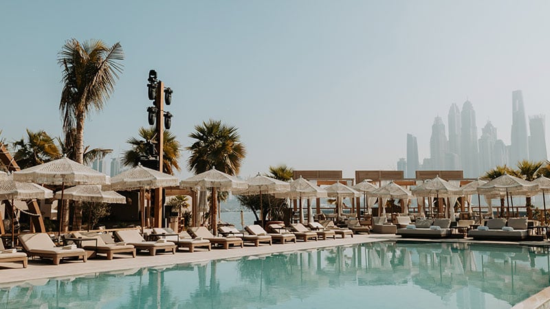 Luxury poolside area with sun loungers and umbrellas, surrounded by palm trees, with a city skyline visible in the hazy background.