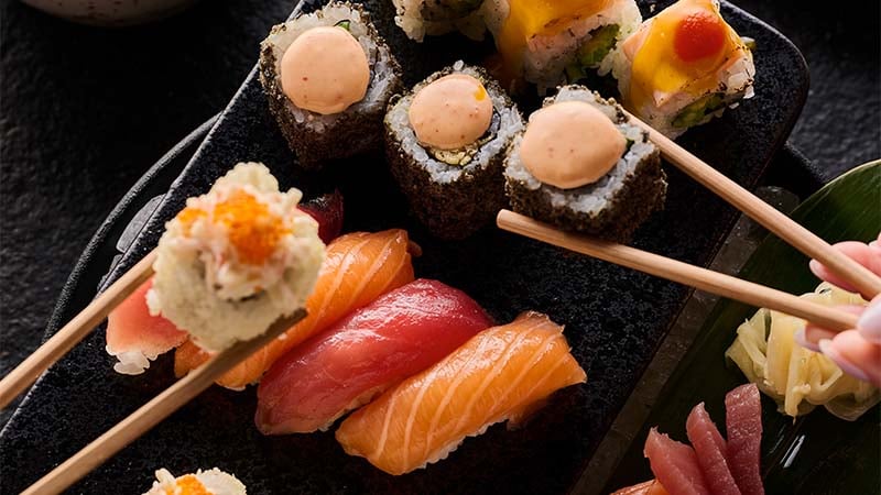 A close-up of assorted sushi rolls and nigiri being picked up with chopsticks from a black serving platter.